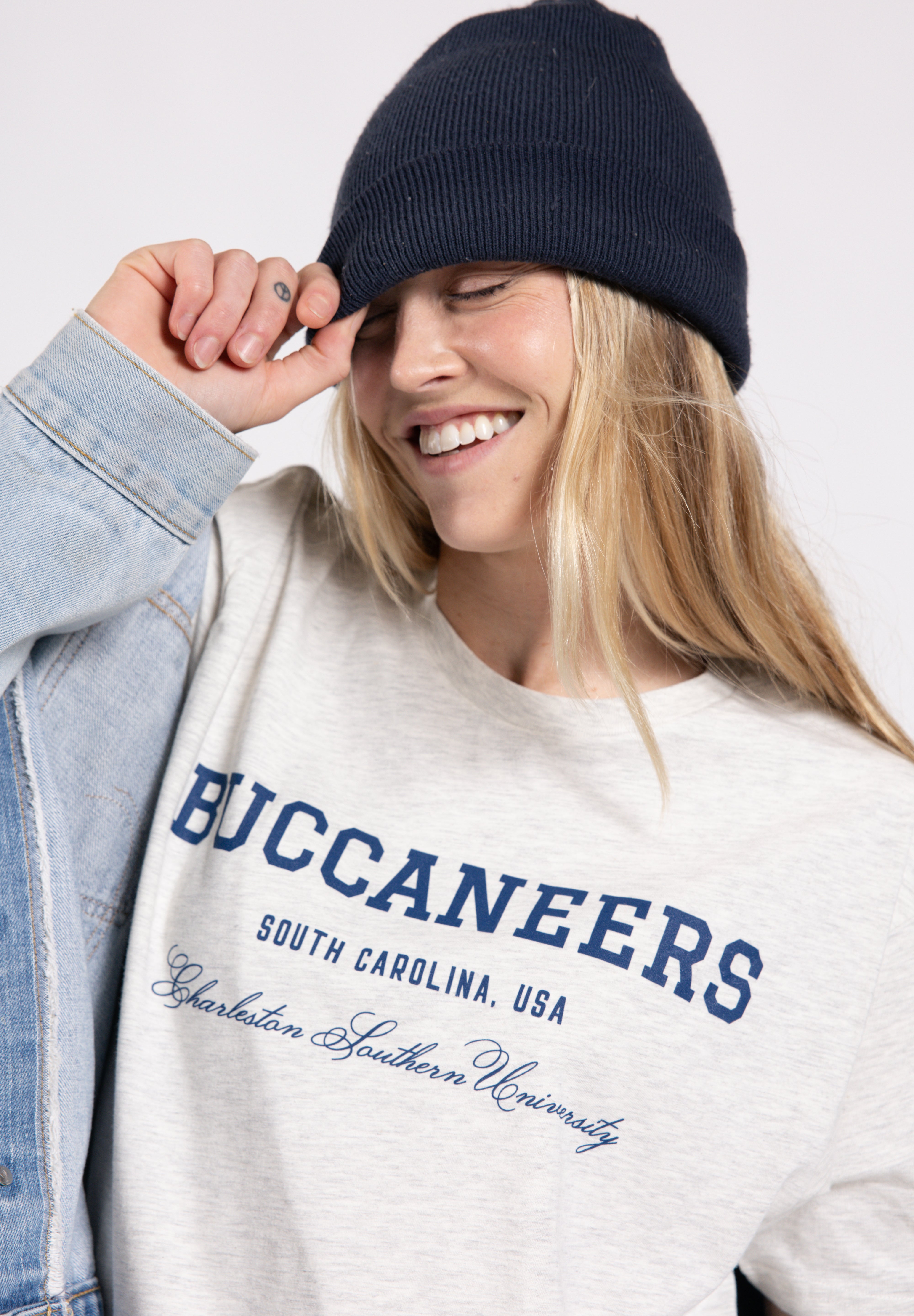 Person wearing a white sweatshirt with 'Buccaneers' text, blue beanie, and denim jacket on a light gray background