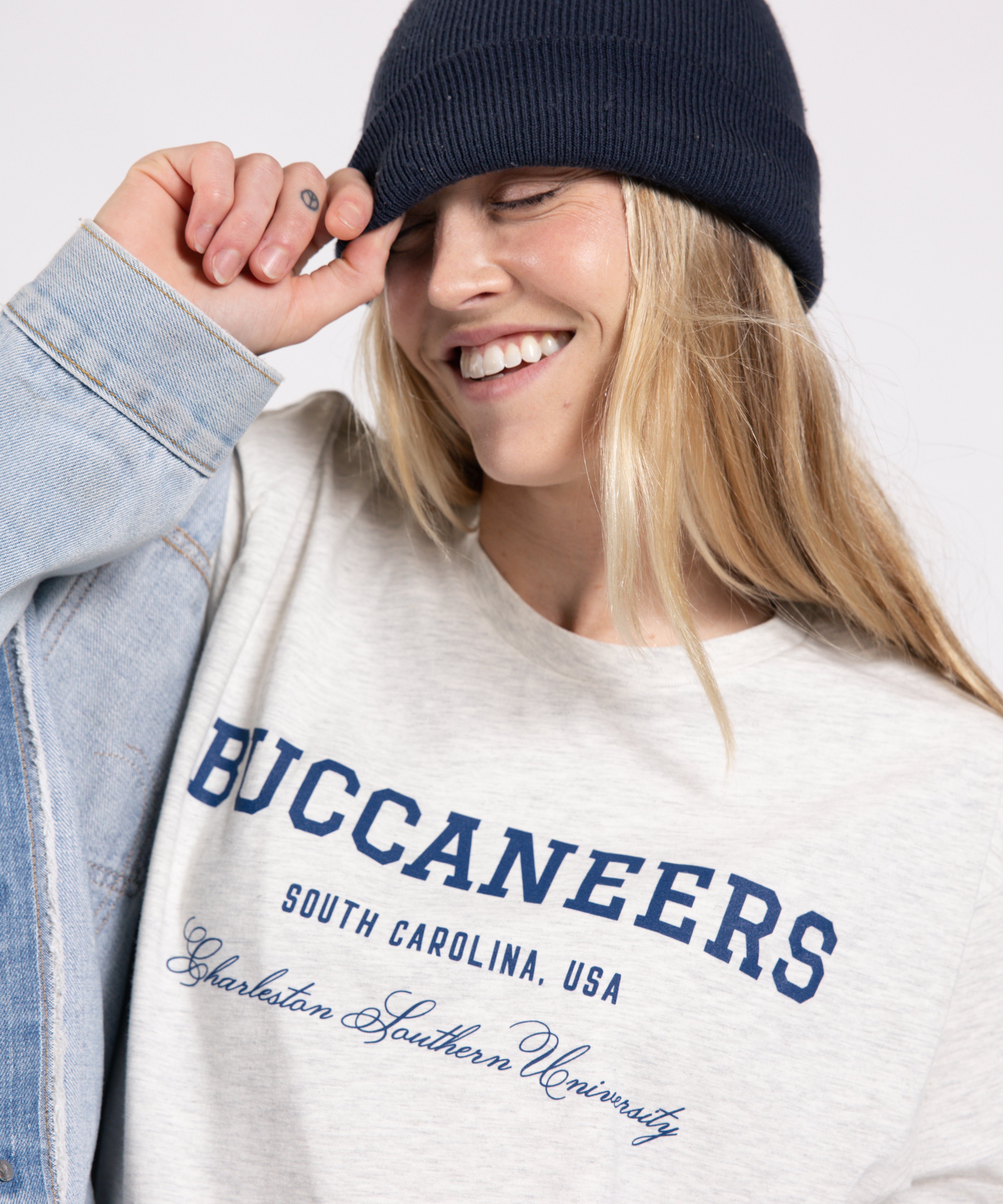 Person wearing a white sweatshirt with 'Buccaneers' text, blue beanie, and denim jacket on a light gray background
